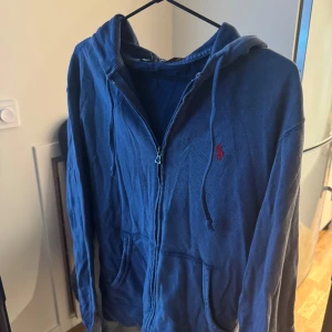 Blå hoodie från Polo Ralph Lauren - Säljer en snygg blå hoodie från Polo Ralph Lauren med dragkedja och huva. Den har en röd broderad logga på bröstet och praktiska fickor framtill. Perfekt för en avslappnad stil.
