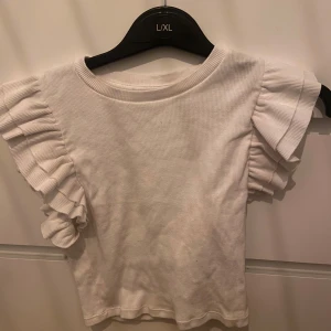 Vit ribbad topp från Zara - Säljer en söt vit ribbad topp från Zara med volangdetaljer på ärmarna. Perfekt för en stilren look. Toppen har en rund halsringning och korta ärmar. Använd en gång!