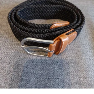 Svart flätat bälte med bruna detaljer - Snyggt svart flätat bälte med bruna läderdetaljer och silverfärgat spänne. Perfekt för att ge en stilren touch till din outfit. Bältet har en flexibel design tack vare det flätade materialet.