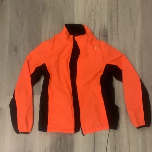 Orange och svart vindjacka - Säljer en sportig orange och svart vindjacka med dragkedja. Perfekt för utomhusaktiviteter med sin lätta och funktionella design. Jackan har långa ärmar och en hög krage för extra skydd mot väder och vind.*Har fläckar se bild 
