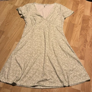 Blommig klänning från NLY - Säljer en söt blommig klänning från NLY i en ljusgrön nyans med vita blommor. Klänningen har en v-ringad design och korta ärmar, perfekt för vår och sommar. Den är gjord i ett mjukt och bekvämt material.