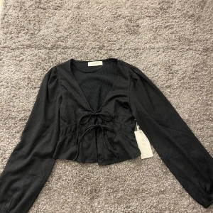 Svart knytblus  - Snygg svart blus från med knytning framtill och v-ringad design. Blusen har långa ärmar och är croppad, vilket ger en trendig look.