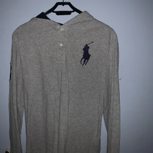 Grå hoodie från Ralph Lauren - Säljer en stilren grå hoodie från POLO med det klassiska logotypen på bröstet. Tröjan har långa ärmar och en knappdetalj vid halsen. Perfekt för en avslappnad look.