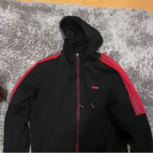 Svart hoodie från Boss - Snygg svart hoodie från Boss med röda detaljer och dragkedja. Den har en huva med snörning och långa ärmar med röda ränder längs axlarna. Perfekt för en casual look. Köpte den på Plick nyligen men säljer den då den var för stor