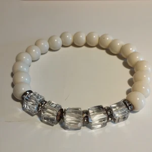 Vitt armband med klara pärlor - Elegant armband med vita runda pärlor och klara, facetterade pärlor som ger en glittrig effekt. Perfekt för att lägga till en touch av stil till din outfit. Armbandet är 6 cm i diameter och cirka 16 cm i omkrets.