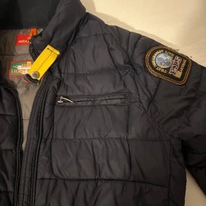 Parajumper Jacka - Säljer en svart quiltad jacka från Parajumpers med en broderad patch på ärmen. Jackan har en dragkedja framtill och en bröstficka med dragkedja. Den är superlätt och perfekt för kyligare dagar.