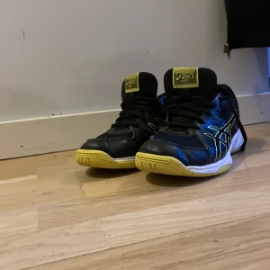 Svarta och gula Asics sneakers - Säljer ett par svarta Asics sneakers med gula detaljer och sula. Skorna har snörning och är perfekta för träning. De har en sportig design med Asics-loggan i gult på sidan.