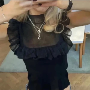 Svart transparent topp med volangdetaljer - Snygg svart transparent topp med volangdetaljer runt axlarna och bröstet. Perfekt för en kväll ute eller en speciell tillställning. Toppen har en elegant och modern design som ger en chic look. Öppen för prisförslag