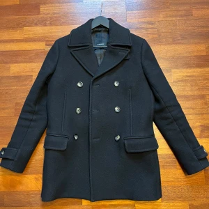 Dubbelknäppt J. Lindeberg rock - Snygg svart rock från J. Lindeberg med dubbelknäppning och stilrena knappar. Jackan har långa ärmar och en klassisk krage. Perfekt för en elegant look.