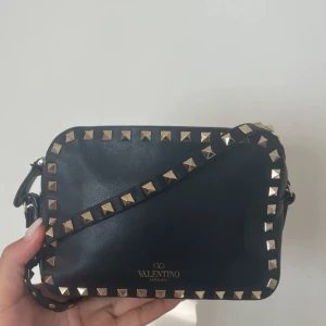 Svart axelväska från Valentino -  Intressekoll på min fina valentino garavani Rockstud camerabag. Äkta men har ej kvitto. Tror det är en tidigare modell med tunnare läder om jag inte har fel. Varsamt använd. Fint skick men tecken på användning. En stud har lossnat Enkelt att byta om man har rätt stud. Eftersom väskan är i läder är den ömtålig och lite skavanker finns på axelbandet men inget som stör användningen av väskan. Lite smutig innuti men generellt bra skick.