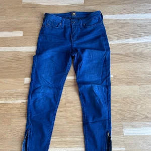 Blå lee jeans Scarlett Cropped - Snygga blå lee jeans med en tight passform och dragkedjor vid bensluten. Modellen är Scarlett Cropped och har en låg midja. 