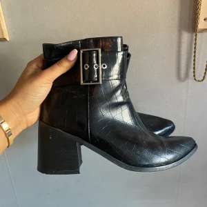 Svarta Boots med Bälte - Snygga svarta boots med en bred bältesdetalj och spänne runt ankeln. De har en spetsig tå och en stabil klack som ger en elegant look.