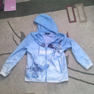 Trapstar windbreaker eagle babyblue - Säljer en blå vindjacka med ett coolt fågeltryck. Jackan har en huva och dragkedja framtill. Perfekt för blåsiga dagar och ger en unik stil med sitt detaljerade mönster.