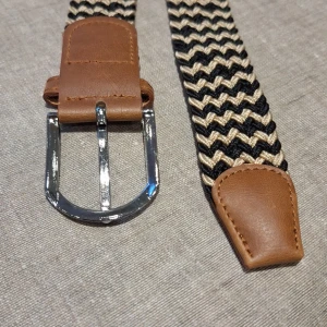 Flätat bälte med bruna läderdetaljer - Snyggt vävt bälte med ett mönster i svart och beige. Bältet har bruna läderdetaljer vid spännet och änden, samt ett silverfärgat metallspänne. Perfekt för att ge en stilren touch till din outfit.