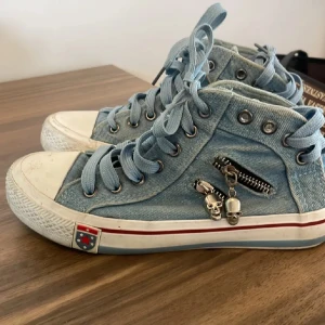 Blå sneakers med döskallar - Coola blå sneakers i jeansmaterial med snörning och unika dödskalle-detaljer vid sidan. Skorna har en vit sula med röd rand och en dekorativ dragkedja. Perfekta för en avslappnad och trendig stil.