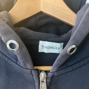 Mörkblå hoodie från Moncler - Säljer en snygg mörkblå hoodie från Moncler med dragkedja och huva. Tröjan har långa ärmar och en klassisk Moncler-logga på ärmen. Perfekt för en stilren och bekväm look.