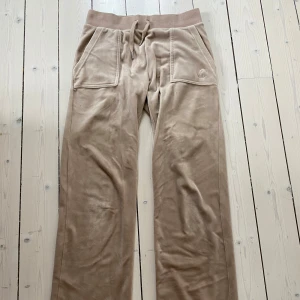 Beige mjukisbyxor från Juicy Couture - Snygga beige mjukisbyxor från Juicy Couture med bred resår och snörning i midjan. Byxorna har fickor både fram och bak, med dekorativa knappar på bakfickorna. Perfekta för en avslappnad stil. Nypris 1400kr säljer för 800kr