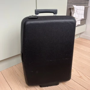 Svart resväska från Samsonite - Säljer en svart resväska från Samsonite med robust design och hjul för enkel transport. Väskan har ett teleskophandtag och ett kombinationslås för säkerhet. Perfekt för resor och enkel att hantera. Inuti finns olika tygfack samt band som håller fast innehållet.  Mått: 55 cm hög, 40 cm lång och 19 cm bred