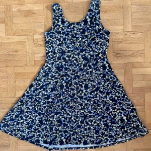 Blommig klänning från Fransa - Säljer en söt blommig klänning från Fransa i mörkblått med ljusa blommor. Klänningen har en normal passform och är ärmlös, perfekt för sommarens varma dagar. Den har en rund halsringning och ett svängigt kjolparti. Finns hund och katt i hemmet.
