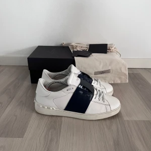 Valentino open  - Snygga vita Valentino sneakers med en bred marinblå rem över ovansidan. Skorna har en unik design med nitar på hälen och är tillverkade i skinn. Perfekta för att ge en stilren touch till din outfit| hör av dig vid minsta fundering 🤔 nypris 7000kr