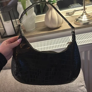 Svart axelväska från Gina Tricot - Snygg svart axelväska från Gina Tricot med krokodilpräglat mönster. Väskan har en praktisk dragkedja och en bekväm axelrem. Perfekt för att ge din outfit en trendig touch. Använd en del men fortfarande fint skick. Minns ej exakta priser men tror runt 300-400kr. 