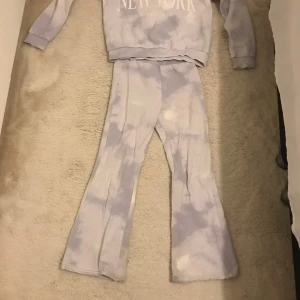 Ljuslila tie-dye set med tröja och byxor - Säljer ett mysigt set i ljuslila och vit tie-dye. Tröjan har texten 'New York' på bröstet och långa ärmar. Byxorna har en utsvängd passform. Perfekt för en avslappnad dag hemma eller en stilren look på stan.