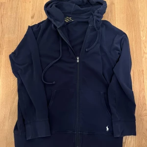 Mörkblå hoodie från Polo Ralph Lauren - Säljer en snygg mörkblå hoodie från Polo Ralph Lauren med dragkedja och huva. Tröjan har långa ärmar och en liten logga på framsidan längst nere. Perfekt för en avslappnad stil.