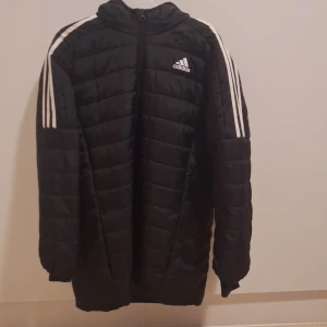Svart dunjacka från Adidas - Säljer en svart dunjacka från Adidas med vita ränder på ärmarna och dragkedja framtill. Jackan har en Luva och är perfekt för kyligare väder. Den har en sportig look med det klassiska Adidas-märket på bröstet.