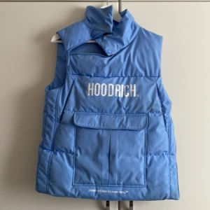 Blå dunväst från Hoodrich - Snygg blå dunväst från Hoodrich med stor logga på bröstet. Västen har hög krage, dragkedja och tryckknappar upptill samt en stor ficka framtill. Perfekt för dig med stil.