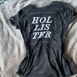 Svart t-shirt från Hollister - Svart t-shirt från Hollister med stort vitt tryck på framsidan där det står 'Hollister Los Angeles California'. T-shirten har rund hals och är kortärmad. Perfekt för en varm sommar dag!