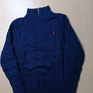 Blå stickad tröja med dragkedja från Polo Ralph Lauren - Mörkblå stickad tröja från Polo Ralph Lauren med halv dragkedja och röd broderad logga på bröstet. Tröjan har kabelstickat mönster och hög krage, perfekt för kyliga dagar.