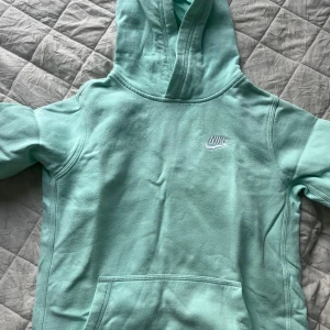 Mintgrön hoodie från Nike - Säljer en mintgrön hoodie från Nike med broderad logga på bröstet. Tröjan har huva och en stor magficka framtill. Perfekt för en avslappnad stil och enkel att matcha med jeans eller mjukisbyxor.