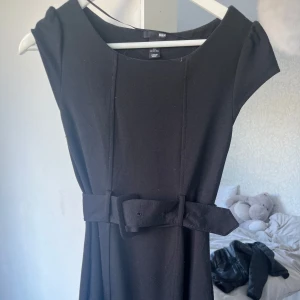 Svart klänning med bälte från H&M - Säljer en svart klänning från H&M med markerad midja och matchande bälte med rosettdetalj. Klänningen har en klassisk, enkel design med rund halsringning och är ärmlös. Perfekt för dig som gillar stilrena plagg.