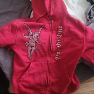 Röd hoodie från One of One med ängelmotiv - Säljer en röd hoodie från One of One med dragkedja. På framsidan finns ett stort ängelmotiv och texten 'One of One' i glittriga stenar. Hoodien har fickor och huva, perfekt för en cool streetstyle-look.