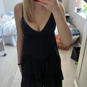 Svart volangklänning med v-ringning - Säljer en svart klänning med djup v-ringning och smala axelband. Klänningen har volangdetaljer och en luftig passform, perfekt för varma dagar. Enkel och stilren design som passar till många tillfällen.