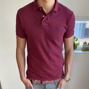 Vinröd Ralph Lauren piké - Säljer en fet vinröd pikétröja från Polo Ralph Lauren (knappast använd) med klassisk krage och två knappar. Tröjan har en broderad blå logga på bröstet och är tillverkad i bomull. Perfekt för en stilren och avslappnad look.