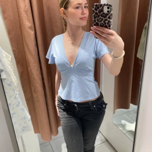 Ljusblå v-ringad blus med volangärmar - Säljer en ljusblå blus med djup v-ringning och korta volangärmar. Toppen har en figurnära passform upptill och faller fint över midjan. Perfekt för dig som vill ha en feminin och stilren look.