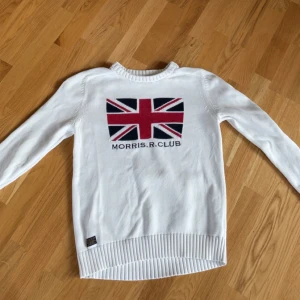 Vit stickad tröja med brittisk flagga från Morris R. Club - Vit stickad tröja från Morris R. Club med brittisk flagga och text framtill. Tröjan har rund halsringning och ribbade muddar vid ärmslut och nederkant. Perfekt för dig som gillar klassisk stil.