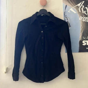 Svart skjorta med knappar - Säljer en svart skjorta med lång ärm och klassisk krage. Skjortan har en figurnära passform och knäppning framtill med svarta knappar. Perfekt att styla till olika outfits.