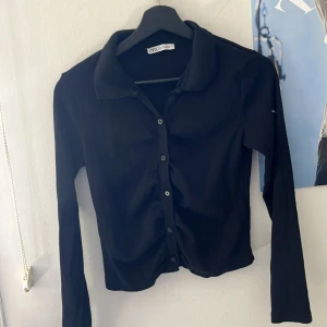 Svart ribbad kofta från Zara - Säljer en svart ribbad kofta från Zara med krage och knappar framtill. Tröjan har lång ärm och är croppad i modellen, perfekt att styla till olika outfits.