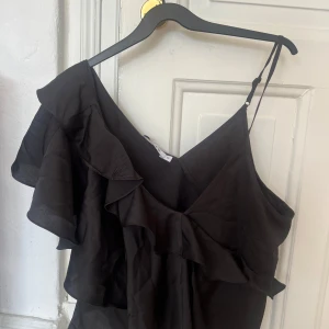 Svart volang topp från NA-KD - Säljer en svart topp från NA-KD med volangdetaljer och en axelbandslös design på ena sidan. Toppen har en asymmetrisk form och tunna axelband på ena axeln. Endast använd ett fåtal gånger, så gott som nyskick. Hör av dig för fler bilder eller frågor!