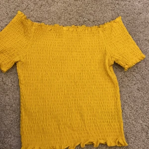 Gul offshoulder topp - Säljer en solgul offshoulder topp med smockad struktur och volangkant. Toppen är elastisk och sitter snyggt på axlarna, perfekt för varma dagar.