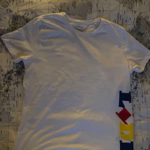 Vit t-shirt med Lidl-logga - Vit t-shirt från Livergy med stor färgglad Lidl-logga i blått, rött och gult längs sidan framtill och en mindre logga på nacken. Klassisk rund hals och korta ärmar. 