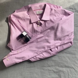 Ljuslila/Rosa randig skjorta från Eton - Ljuslila/Rosa Randig Skjorta från Eton.        Storlek: Slim 42.      Säljer en stilren ljuslila skjorta från Eton i slim fit med extra långa ärmar. Skjortan har diskreta vita ränder, klassisk krage och vita knappar. Perfekt för dig som vill ha en dressad look med lite färg.