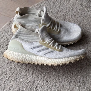 Vita Adidas Ultraboost sneakers - Säljer ett par vita Adidas Ultraboost sneakers med stickad ovandel och högt skaft. Skorna har snörning, platt sula och diskreta ränder på sidan. Perfekta för dig som gillar stilrena och bekväma skor.