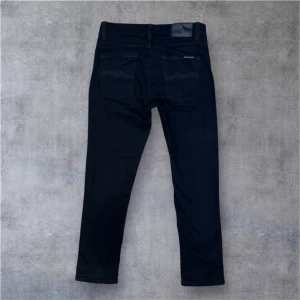 Svarta jeans från Nudie Jeans - Säljer ett par svarta jeans från Nudie Jeans med klassisk femficksdesign och raka ben. Byxorna har silverfärgade nitar och knappar samt diskret logga på bakfickan. Perfekta för en stilren look. Perfekt för sommaren! Priset inte hugget i sten och hör av dig vid frågor 