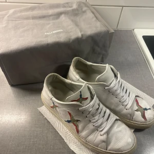 Vita sneakers från Axel Arigato med broderade fåglar - Säljer ett par vita sneakers från Axel Arigato med färgglada broderade fåglar på sidorna och framtill. Skorna har snörning och platt sula. Perfekta för dig som gillar stilrena men unika detaljer. Lite värre skick nu då 