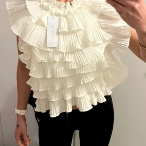 Vit volangblus med plisserade detaljer från Feelhoo - Supersnygg vit blus med flera lager volanger och plisserade detaljer. Hög halsringning med rynk och breda volangärmar. Från Feelhoo men ser exakt ut som Le magda blusen. Storlek m/l men passar mig som är S