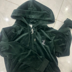 Mörkgrön velour hoodie från Juicy Couture - Mörkgrön hoodie i mjuk velour från Juicy Couture med dragkedja och huva. Stenprydd Juicy-logga på ryggen och ett glittrigt emblem på bröstet. Perfekt för en avslappnad och trendig look.