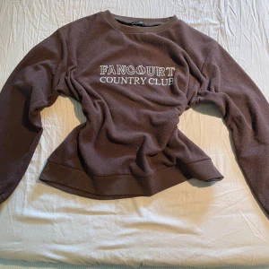 Brun sweatshirt från SHEIN med text - Mysig brun sweatshirt från SHEIN med texten 'Fancourt Country Club' broderad på bröstet. Tröjan har långa ärmar, ribbade muddar och är tillverkad i ett mjukt syntetmaterial. Perfekt för en avslappnad stil.